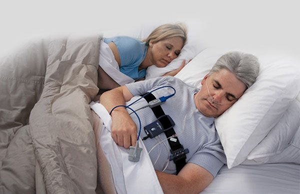 Man and woman sleeping in bed; man wears resmed sleep test monitoring device. Beige bedding, calm atmosphere. No visible text.