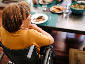Woman in a wheelchair at a dining table with plates, salad in a blue bowl, and wooden floor. She's wearing a yellow shirt, looking content.