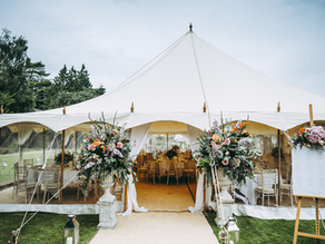 White marquee with floral arrangements stands on a grassy lawn. Inside, tables and chairs are set up, visible through clear sides. A seating chart is on display.