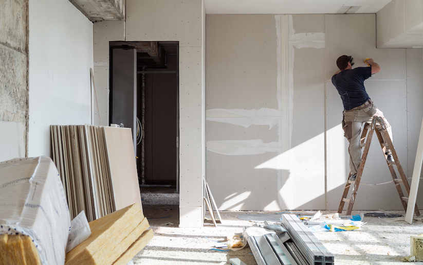 Worker on ladder installs drywall to install wiring in a sunlit room with scattered construction materials. Walls are partially spackled; bright setting.