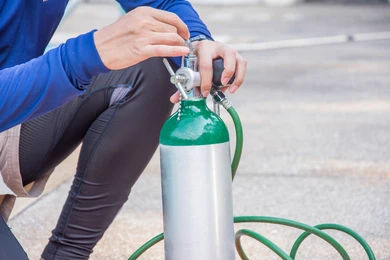 Person in blue adjusts a green and silver oxygen tank outdoors. Green hose is visible on a concrete surface, creating a focused, calm scene.