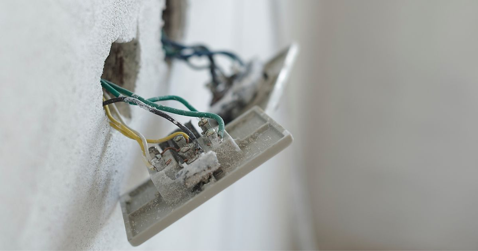 Exposed electrical wiring hang from a wall socket in a white room. The wires are green, yellow, and blue, and the socket is dusty.