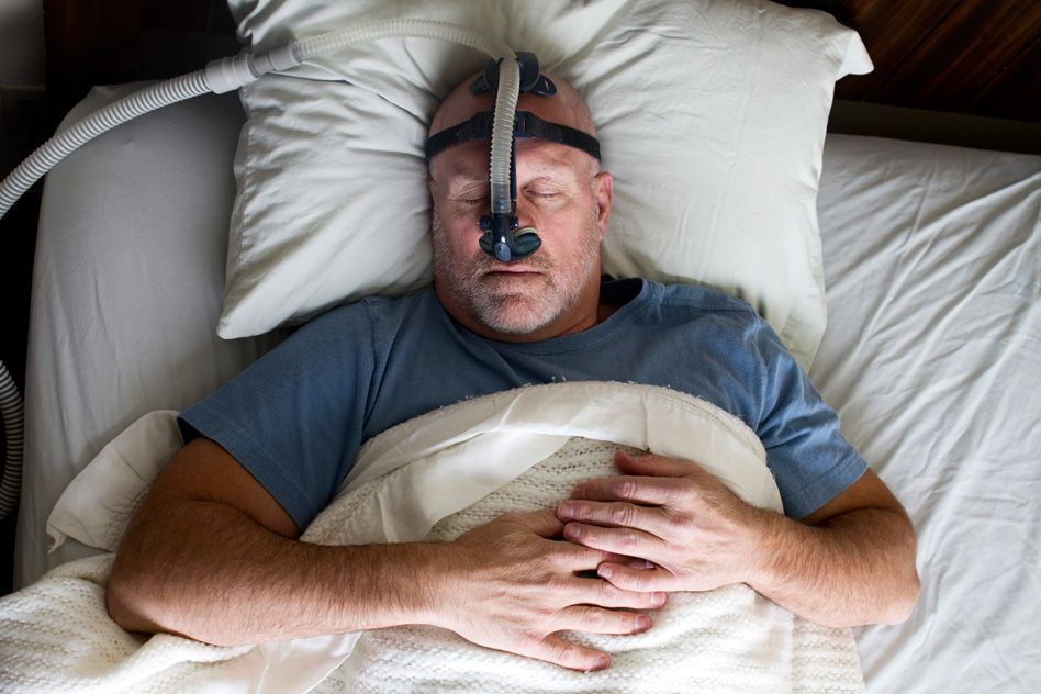 Man sleeping in bed wearing a CPAP mask, hands resting on chest. White sheets, blue shirt. Calm and restful atmosphere.