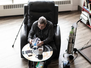 Man sitting in chair assembling a CPAP machine, with a cup, phone, and books on the table. Oxygen tank and crutch beside him. Light setting.