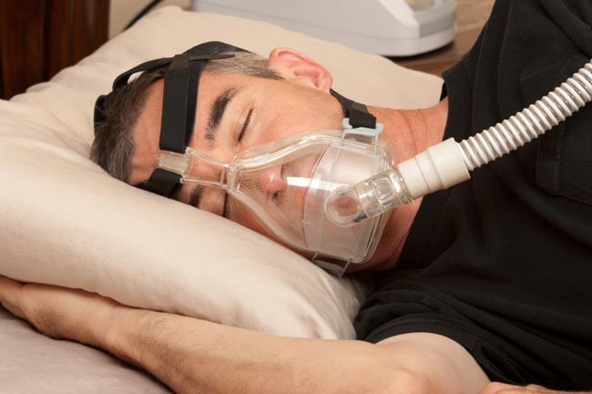 Lelaki tidur dengan topeng CPAP, berbaring di atas bantal kuning air di dalam bilik tidur. Pemandangan itu tenang, dengan tiub jernih topeng kelihatan.