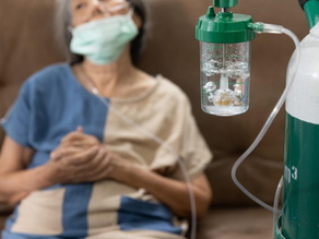 Elderly person with a mask sits on a couch, connected to a green and white oxygen tank. Blurred background conveys a calm mood.