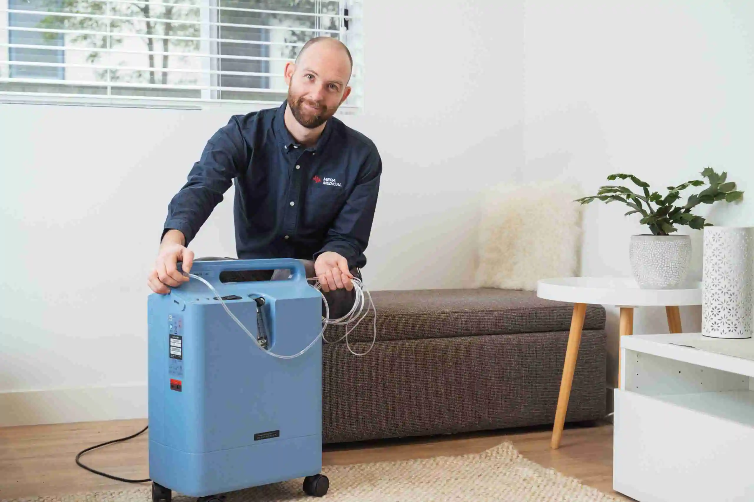shows a man sitting on a couch, in a bright room with a rented oxygen concentrator, in blue.