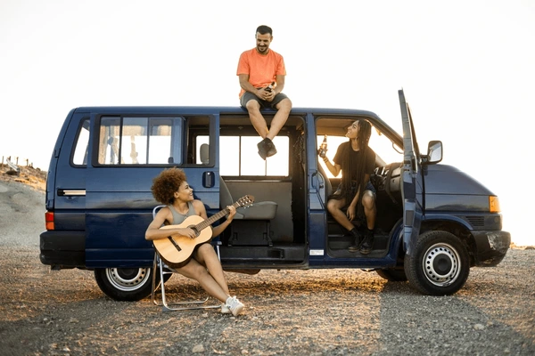 Tiga orang bersama van biru di kawasan berbatu ketika matahari terbenam. Seorang bermain gitar, seorang lagi duduk di atas van, dan seorang lagi berada di dalam, memegang minuman. Suasana santai.