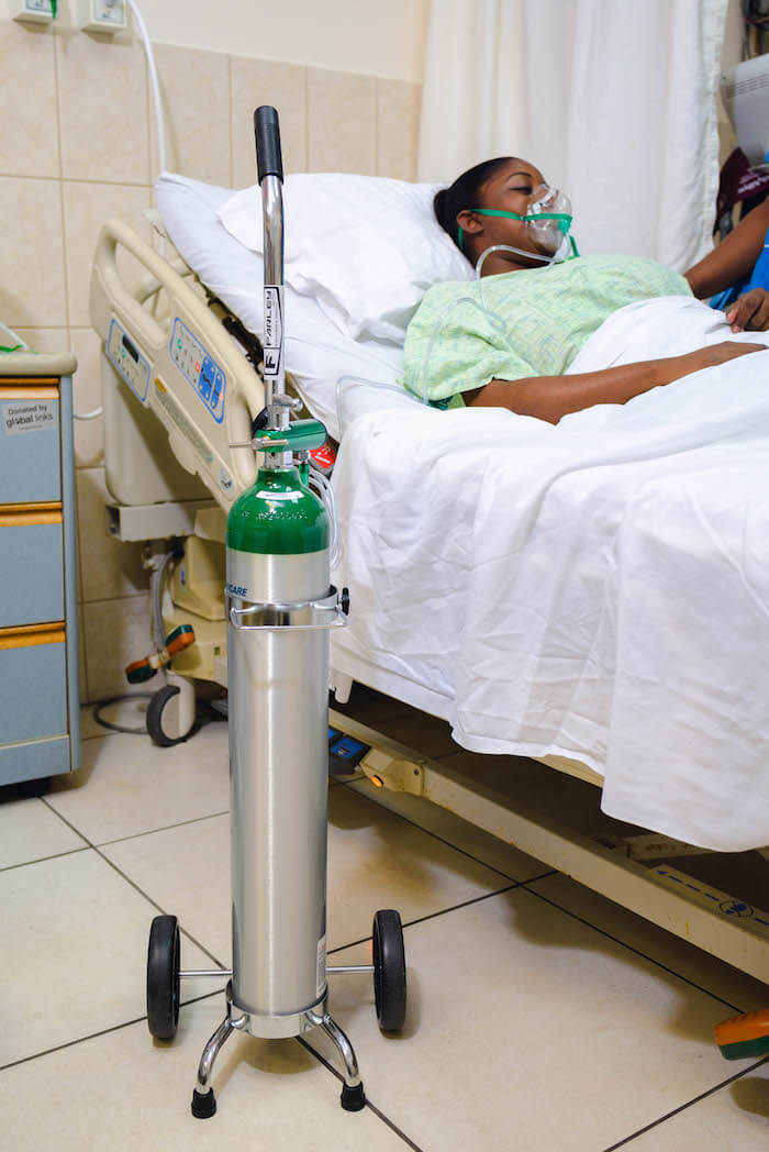 Seorang pesakit berbaring di atas katil hospital dengan topeng oksigen, disambungkan ke tangki oksigen hijau dan perak di atas roda. Tetapan hospital.