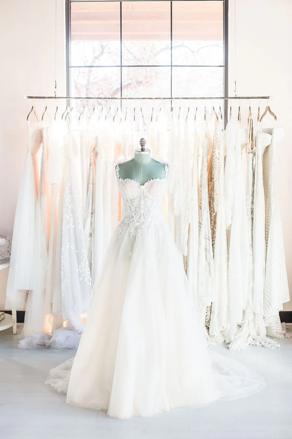 A white wedding dress on a mannequin stands in front of a rack of dresses in a bright room with a large window, creating a serene atmosphere.