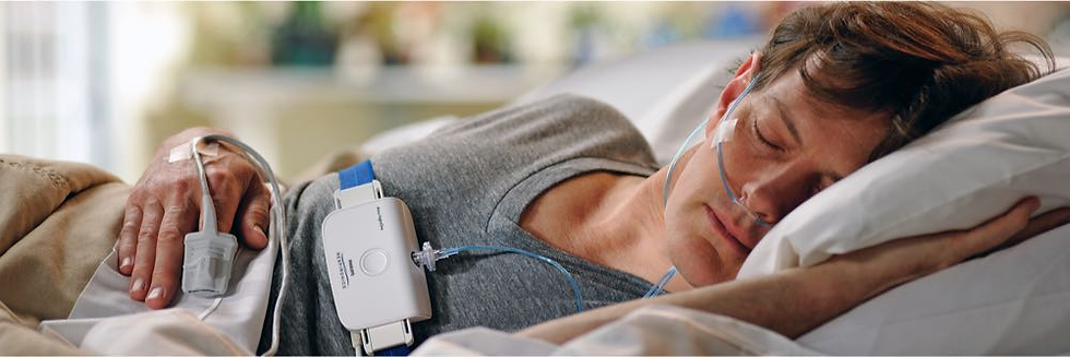 A person sleeps in a bed with sensors attached. They appear calm. The room is softly lit, and a device with visible text is on their chest.