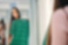 Woman in a fitting room holds a green dress, looking at her reflection in a mirror. Background has blue curtains. Calm, contemplative mood.