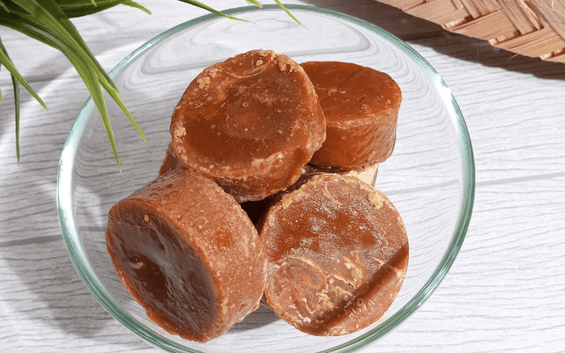 Brown sugar discs stacked in a glass bowl on a light wooden table. Green plant leaves in the corner, wicker mat visible in the background.