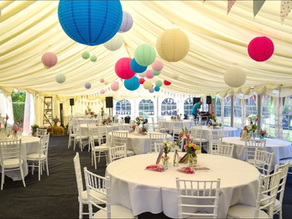 White tented venue with colorful paper lanterns and bunting. Round tables with white chairs, floral centerpieces. Vibrant, festive mood.