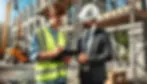 Two construction workers, one in a yellow vest writing on a clipboard, and another in a suit with a hard hat, discuss at a building site.
