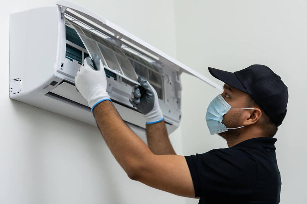 Person in gloves and mask servicing air conditioner on a white wall. Wears black cap and shirt, focused and carefully working.
