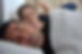 Man sleeping under a white blanket, mouth open. Background blur shows a woman looking annoyed. Indoor setting with neutral tones.