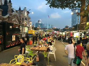 Crowded street food market with food trucks, tables, and people dining outdoors. Historic building and cityscape in the background.
