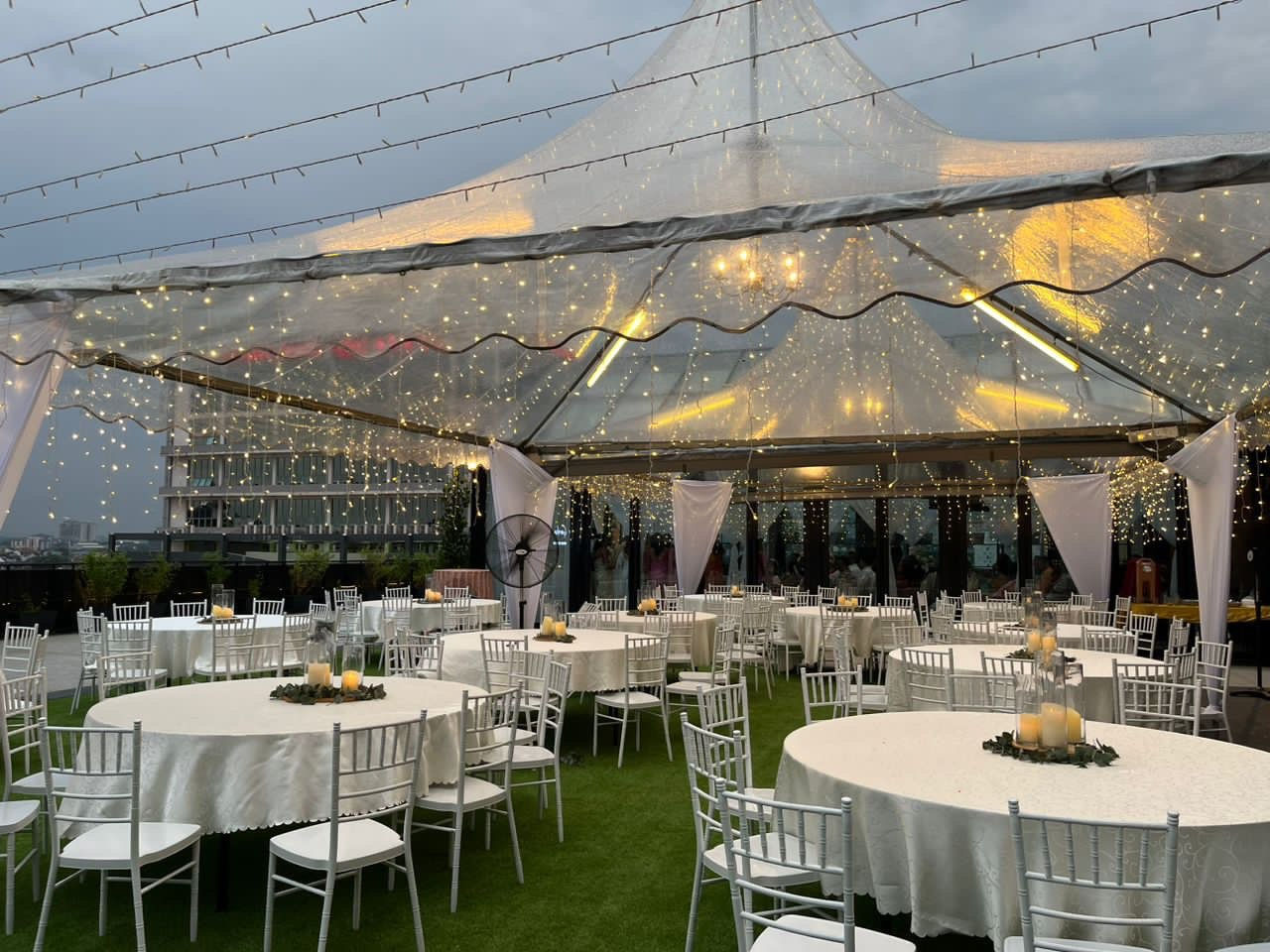 Luxurious event under a transparent tent with rented white chairs and tables and decorative chandeliers.