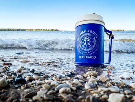 Well-worn blue and white plastic coffee mug on a beach with the Outward Bound logo.