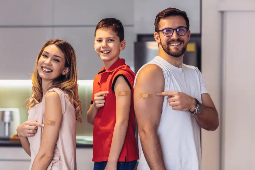 A family smiling and pointing to bandaids on their arms after receiving vaccinations.