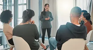 A young woman stands in front of a group of people sitting in a circle during a group ther
