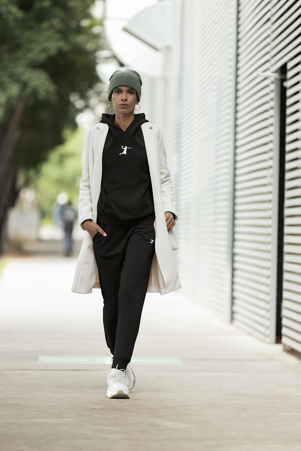 Modèle portant le jogger Player Shadow noir avec logo blanc, manteau long blanc et casquette noire, marchant en extérieur