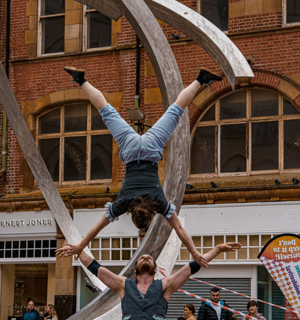 One performer is upside down above the other perfomers, and they are looking at each other. 
Their arms and legs are out to the sides. 
They are performing in front of a metal sculpture. 