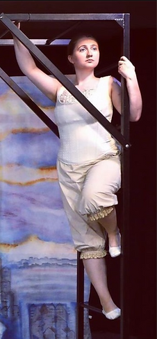 Calista Hoyer, in a white corset and bloomers, holds herself upright on the side of a jungle-gym frame, looking out stoically into the audience.