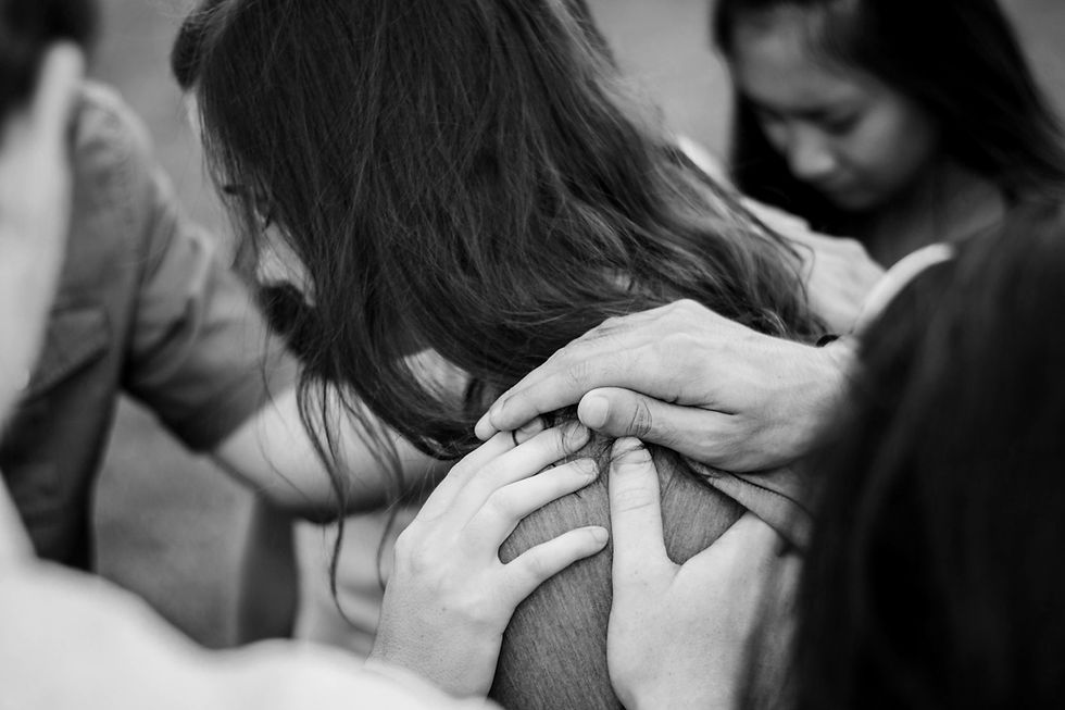 Black-and-white close-up of several people resting hands on a woman’s shoulders in a supportive group embrace.