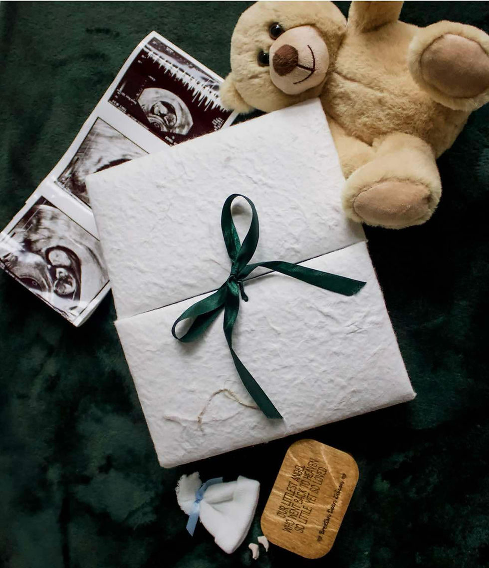 A flat lay featuring a soft beige teddy bear, a folded white textured paper tied with a dark green ribbon, and a strip of black-and-white ultrasound images partially tucked under the paper. The scene also includes a small white fabric pouch with a blue ribbon and a wooden plaque engraved with a heartfelt message. All items are placed on a dark green, plush fabric background.