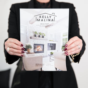 Woman holds Kelly Malina Realtors brochure with home interior design displayed.