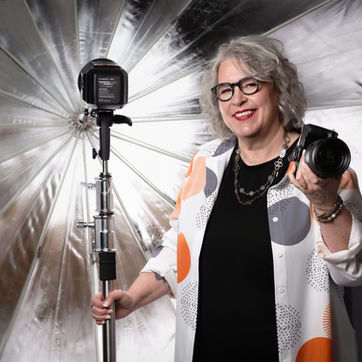 Smiling woman holding camera, wearing glasses, in front of photography equipment 