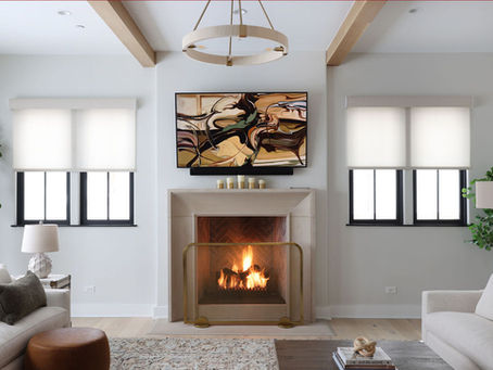 Modern living room with a lit fireplace below abstract art. Neutral tones with beige sofas, plants, and large windows creating a cozy atmosphere.