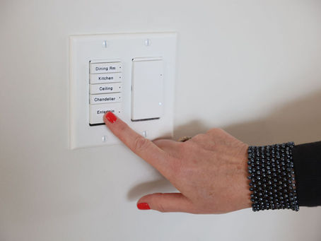 Hand with red nail polish presses “Entertain” button on a white light switch panel. Beaded bracelet visible, set against a light wall.