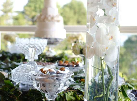 Um ramo de orquídeas brancas em um cilindro de vidro a frente, com o bolo da festa ao fundo