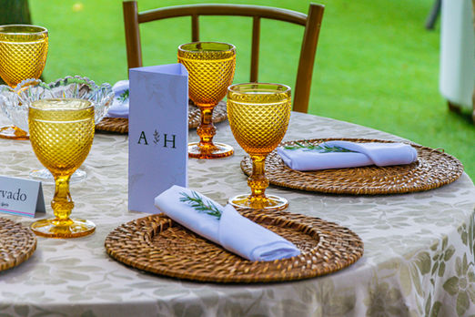 Um sousplat de vime, combinando com a taça em marrom e um guardanapo de tecido branco com um pequeno raminho de alecrim, montados numa mesa redonda com toalha em ocre