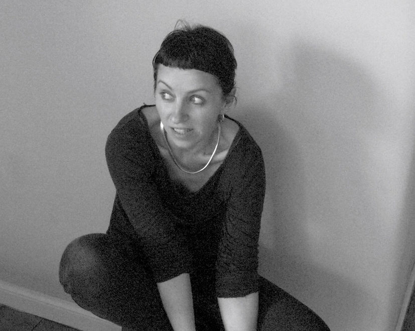 a black and white photograph of me sitting on a stool listening and attentive. I am a white woman and I have short dark hair, a nose ring and am wearing a dark t-shirt and jeans.