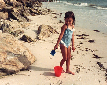 A young child wearing a blue and white gingham swimsuit with ruffled trim stands barefoot on a sandy beach near some large rocks. She holds a small blue plastic shovel in one hand and looks down toward a bright red bucket at her feet, with waves and scattered seaweed visible along the shore in the background.