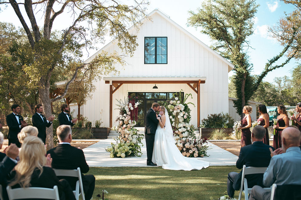 Outdoor wedding ceremony with couple kissing in front of white chapel and floral arch.