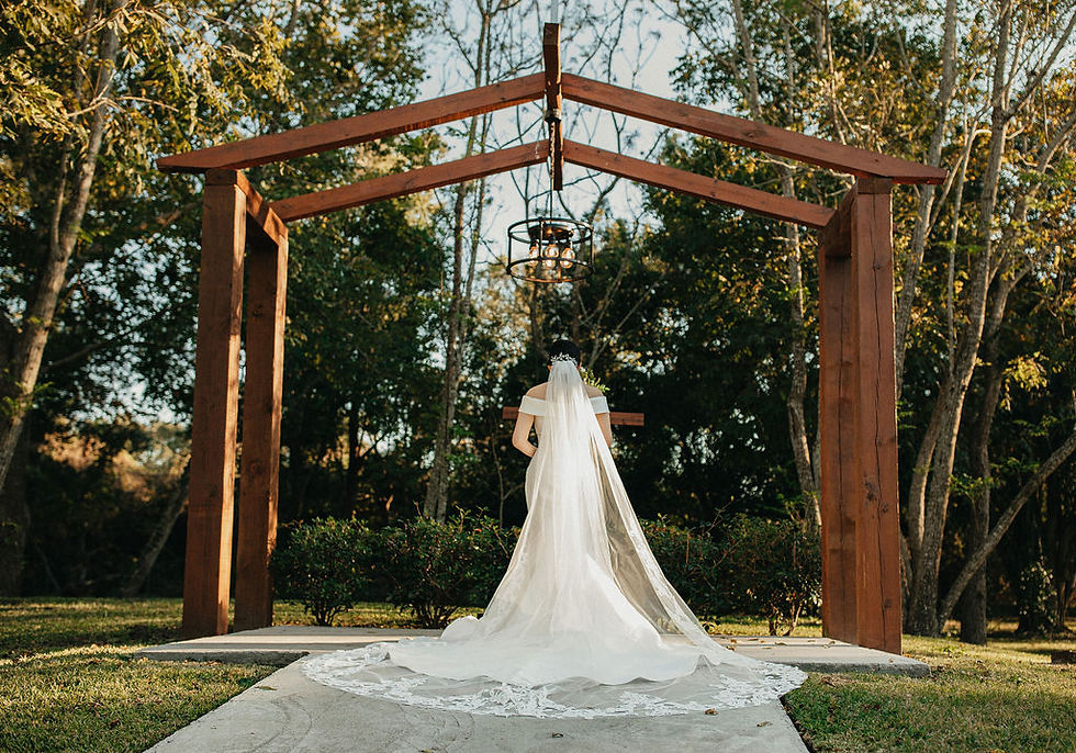 Bride stands beneath wooden arch with long veil at outdoor Houston ceremony.