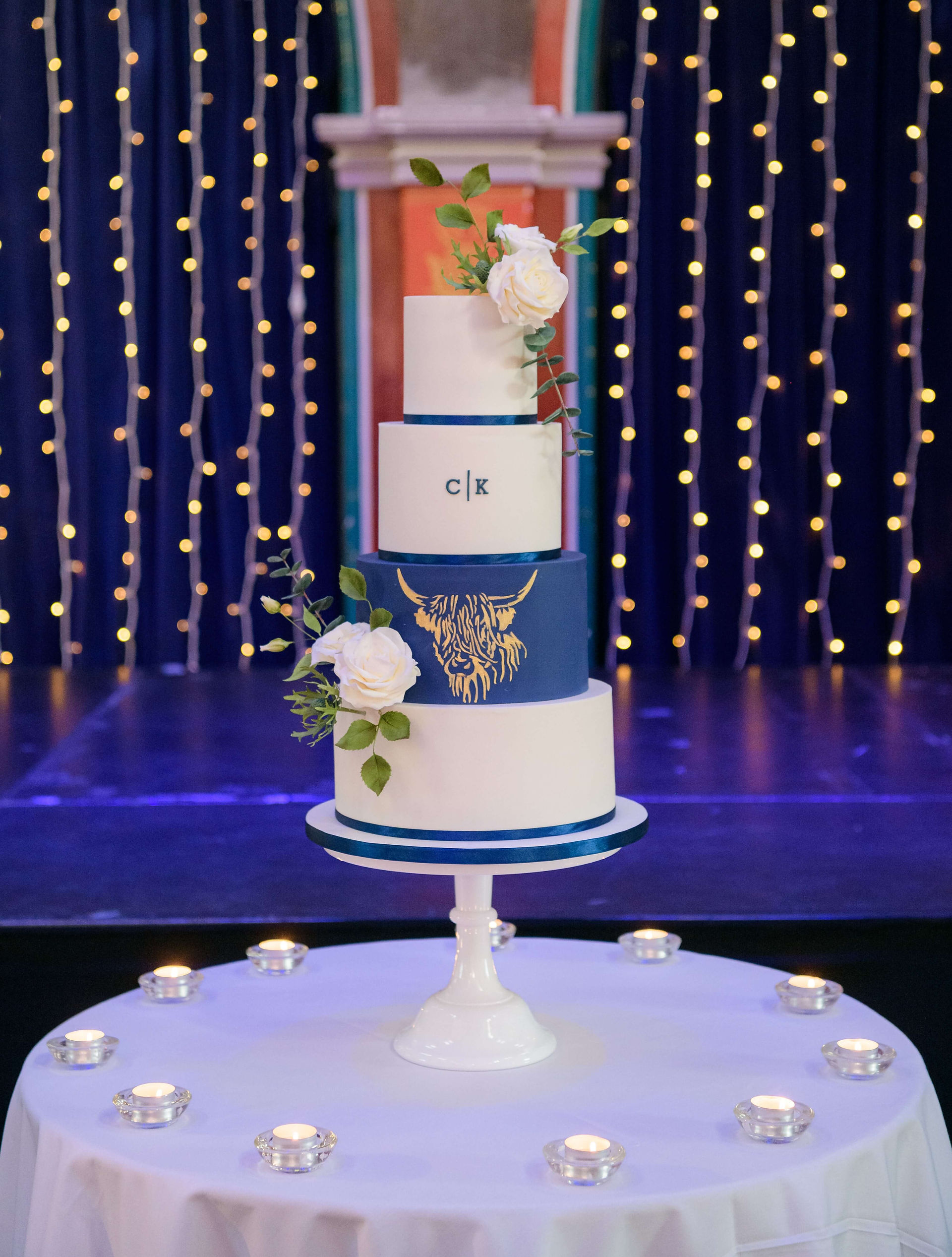 4 tier cake. The 3rd tier is navy and has a gold Highland cow stencil on it. It's on a table surrounded by tealights and has fairy lights in the background