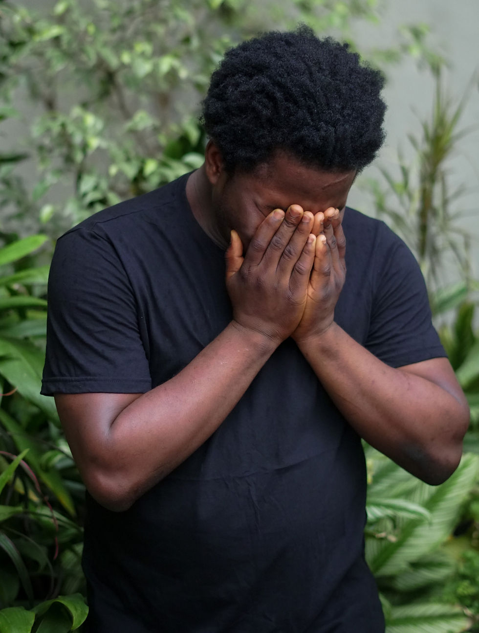 Homem negro com camiseta preta cobrindo o rosto com as mãos mostrando sinais defrustração e cansaço.