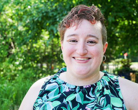 Abby, a white nonbinary person with short curly hair, smiles at the camera. They are wearing a green botanical print dress and small hoop earrings.