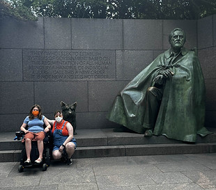 In front of statue at the FDR memorial a young disabled white woman sits in a wheelchair with a disabled nonbinary white person squatting next to her. 