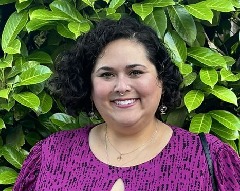 an image of a Mexican, jewish, disabled woman with short curly hair, a septum piercing and a pink and black top. Davi is similing while standing in front of a green bush