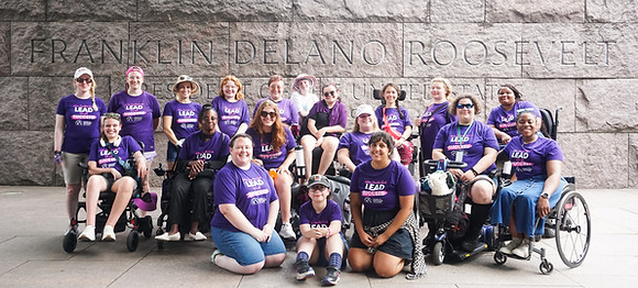 a diverse group of disabled women and nonbinary people wearing purple tshirts posing infront of the FDR memorial sign in DC