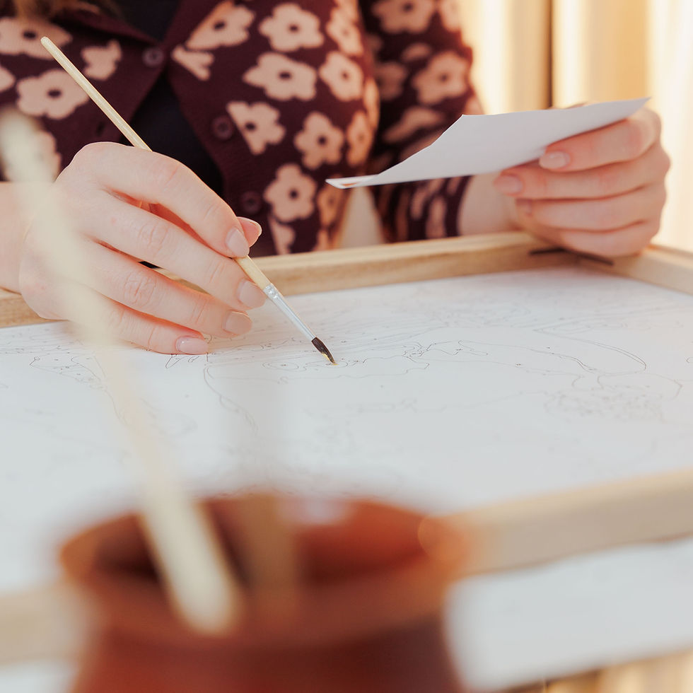 Thumbnail: Close-up of a fine-detail paintbrush on a numbered canvas and a woman holding a reference paper to help guide her painting.