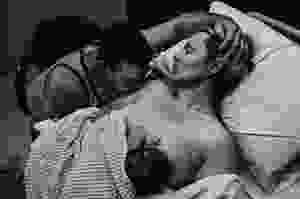 a black and white image of a woman who has just given birth, laying in the hospital bed breastfeeding her newborn. her eyes are closed and her husband sits next to her, stroking her head and kissing her arm.