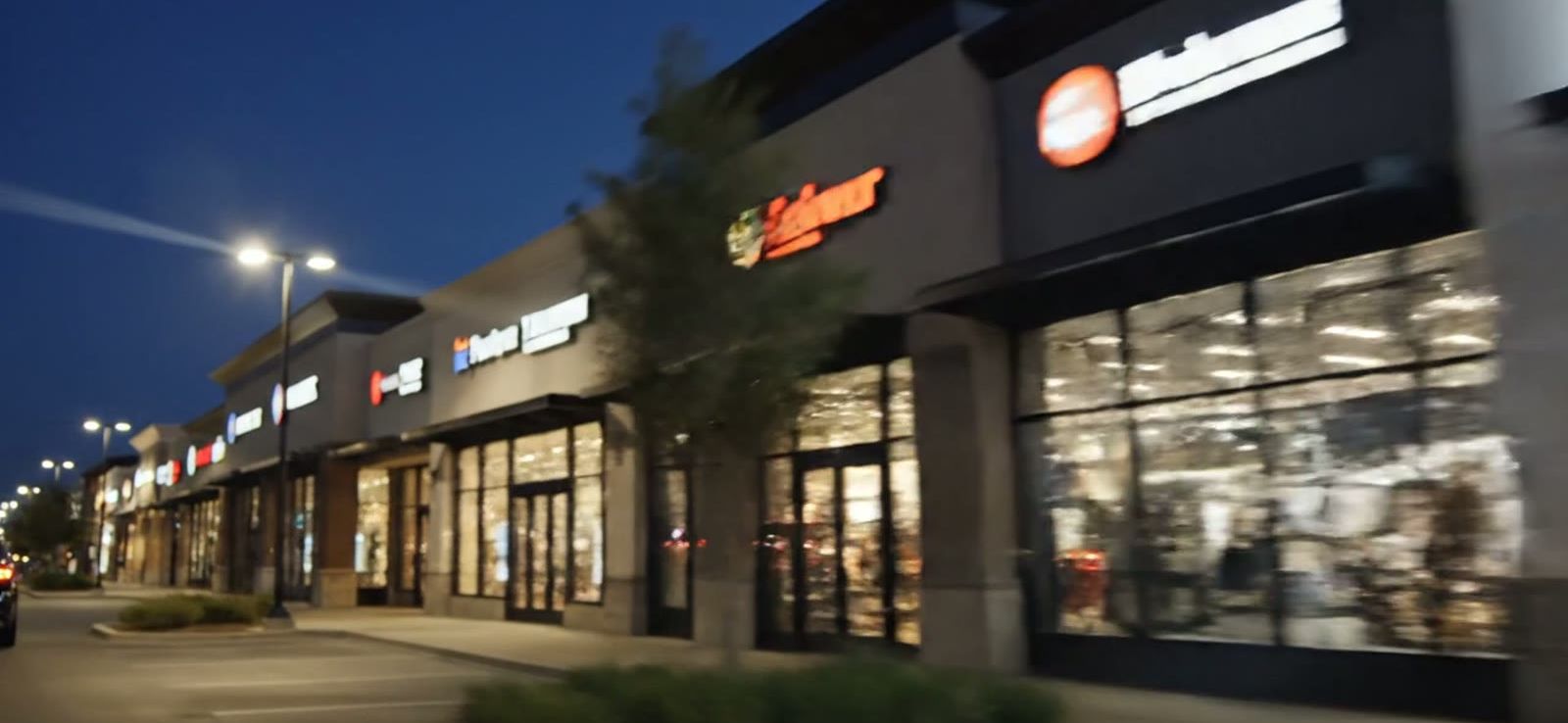Multi-store retail shopping center at dusk with lit storefronts and parking lot lighting